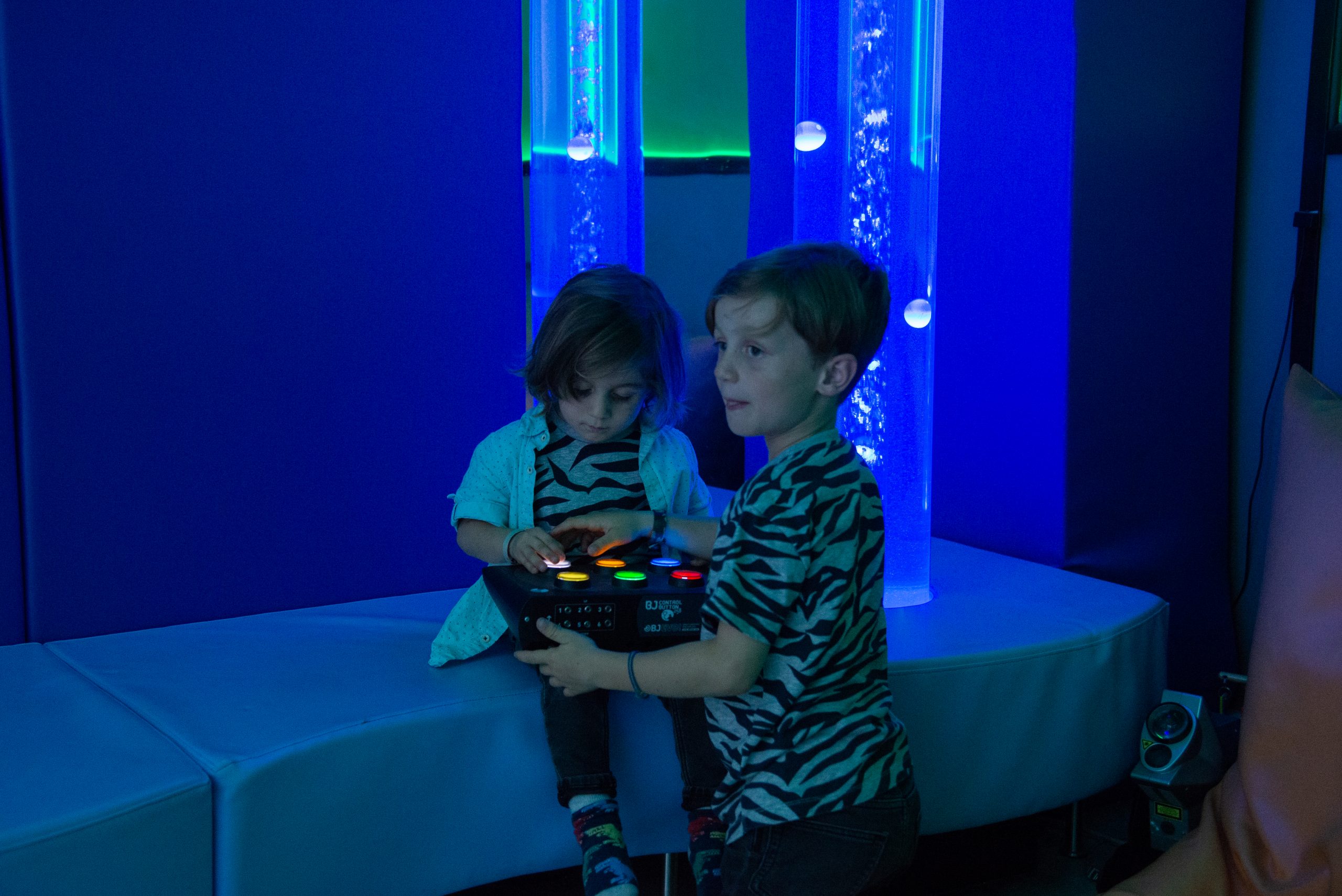 Dos niños utilizando un conmutador de seis botones para interactuar con el tubo de burbujas de la sala multisensorial